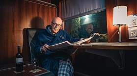 Mann im Nachtmantel liest bei gemütlichem Licht in einem Schlafwagen eine Zeitung