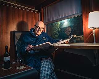 Mann im Nachtmantel liest bei gemütlichem Licht in einem Schlafwagen eine Zeitung